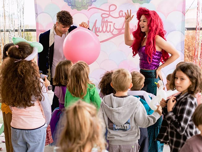 Doi animatori și un grup de copii în jurul unui balon roz mare; fotografie într-un spațiu de petrecere.